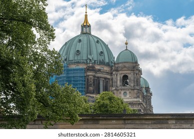 View of Berlin Cathedral with dramatic clouds and lush European oak trees. Historic architecture dominates the skyline with restoration scaffolding visible.
