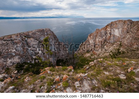 View between two rocks on Lake Baikal from Olkhon Island. On the stones is red moss, near the vets and grass. Mountains behind the lake. Clouds in the sky. There is a dry tree and green.