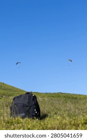 view of a black backpack resting on a grassy green ridge below a vast clear blue sky with two paragliders in the distance and extensive copy space.