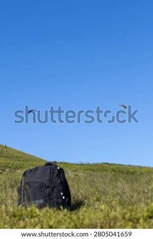 view of a black backpack resting on a grassy green ridge below a vast clear blue sky with two paragliders in the distance and extensive copy space.