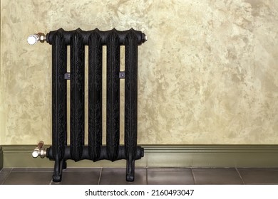 View of a black cast-iron heating radiator in the corner of a room against a beige wall.