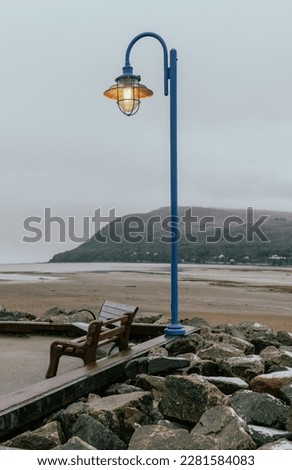 view of a blue metal retro looking lamp post and a bench next to a paved road  during fall on a cloudy day with a mountain behind
