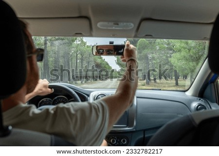 View of a blurred man adjusting rear view mirror while driving on a road through pine forest.