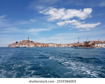 A view from a boat of a coastal town with colorful buildings, a prominent church tower and boats on the blue sea under a bright sky with occasional clouds. The scene exudes charm and tranquility. - Powered by Shutterstock