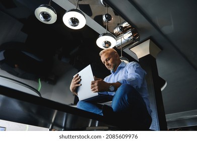 View From The Bottom To A Handsome Middle Aged Caucasian Man In Casual Denim Jeans And Blue Shirt Working On Laptop Sitting On A Table At Home Kitchen, Working Remotely From Home