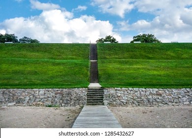 A view from the bottom of the hill while standing on a beach boardwalk. Stone stairs ascend into the sky over a grassy hill. Benches peek out from the top of the hill against a cloudy blue sky.