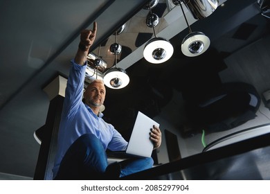 View From The Bottom To A Successful Middle Aged Caucasian Man In Casual Denim Jeans And Blue Shirt With Laptop, Pointing Finger Up, Sitting On A Table At Home Kitchen, Working Remotely From Home