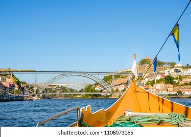 View of a bow of a yellow boat floating down Douro river in Porto, Portugal in the summer; Dom Luís  Bridge and multiple buildings in the background