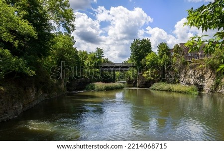 View of bridge over the rapid Grand River, rocky banks, green trees, cloudy sky, day time. Fergus, Ontario, Canada