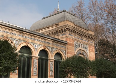 View Of A Building With Archways And Colourful Decorations Partially Obstructed By Trees On The Foreground In El Retiro Public Park In Madrid, Spain.