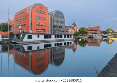 View of the Bydgoszcz waterfront featuring a modern glass building and a historic red brick structure, with reflections in the Brda River in Poland - Powered by Shutterstock