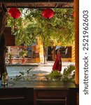 View from a cafe window in Hoi An, Vietnam, capturing a woman in a red dress and traditional conical hat walking down the street. The scene is framed by lush greenery and vibrant red lanterns.