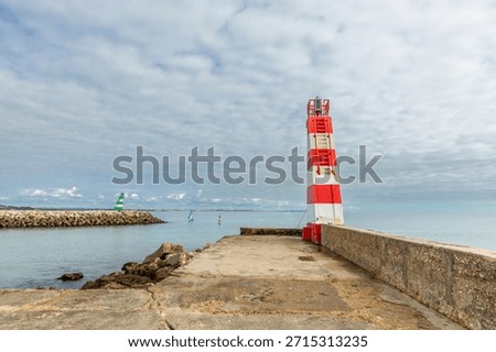 View of the Cais da Solaria lighthouse at Praia da Batata in Lagos, Algarve, Portugal. The red and white lighthouse stands on a concrete pier overlooking calm Atlantic waters, with sailboats.