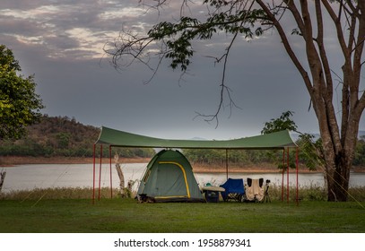 View of a camping tent with awning pitched On the grass under beautiful big trees with branches at the campsite lake shore with mountain range in background.  - Shutterstock ID 1958879341