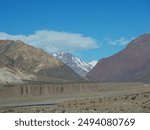 View of a canyon in a desert region with a meltwater river flowing through it, and snow-capped mountains of the Andes in the background under a blue sky during winter in Mendoza, Argentina