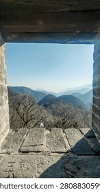 A view captured through the stone window of an ancient Great Wall watchtower reveals layered misty mountains stretching into the horizon under a clear blue sky. The weathered stone frame and wooden beam ceiling highlight the historic architecture, contrasting with the natural beauty of the distant hazy peaks beyond. This scene captures the timeless connection between human history and nature.
