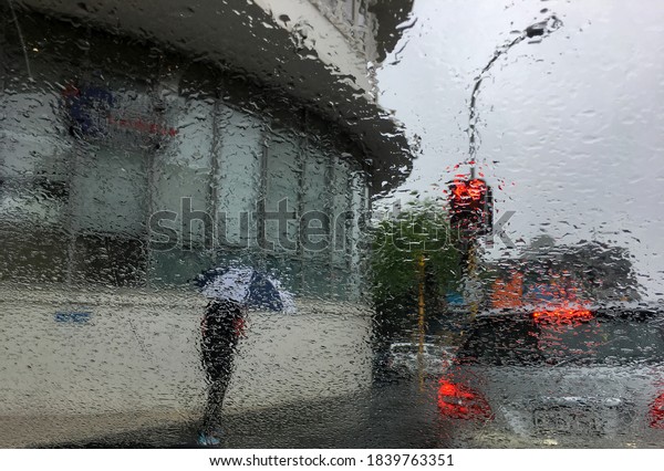 The view from the\
car window of the intersection in the rain. The red traffic light\
is on. Sick raindrops on the windshield blur the image. A man walks\
under an umbrella.