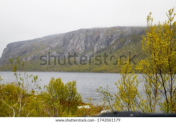 View from the car window of a\
sad, harsh, dramatic landscape with a lake, river or fjord, a rock\
in the distance on the horizon and a cloudy, rainy, gray\
sky