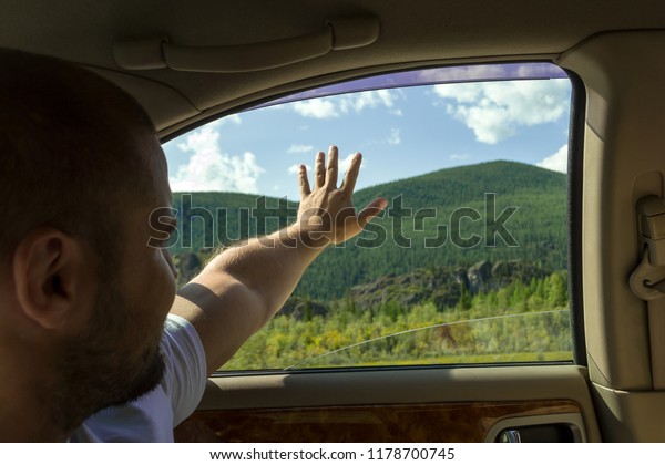 A view from the car\'s interior to the man who sits in\
the back seat and extends a hand in the window saying goodbye or\
greeting the nature and mountains of the Altai rejoicing in\
traveling by car