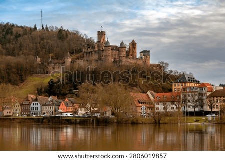 View of the castle from the Main River. A dramatic wide-angle view of the medieval Burg Wertheim perched high on a hill above the Main River valley. The historic stone ruins overlook a vibrant autumn landscape, where forests glow in shades of gold, amber, and red. The winding river reflects the soft evening light, creating a serene and cinematic European countryside scene. A perfect representation of autumn in Germany with rich history and natural beauty.