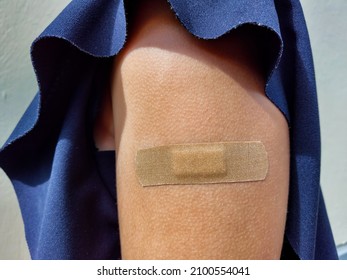 View Of A Caucasian Girl In Dark Blue Blouse With Naked Upper Arm With Brown Bandaid After Getting A Dose Of The COVID-19 Vaccine Outdoors In Bright Sunlight