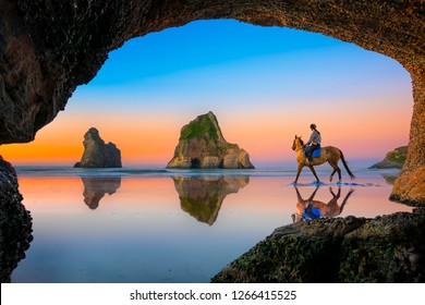 View From The Cave Of Scenery View The Sunset Of Wharariki Beach With Woman Riding Horse On The Reflection Beach, New Zealand Summer Tourist Popular Place For Vacation Long Weekend