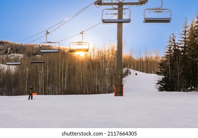 View Of Chairlift In Colorado Ski Resort In Winter Afternoon; Sun Setting In The Woods Behind The Chairlift