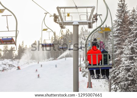 View from chairlift over ski piste, skier in bright red jacket seating in front, more blurred people skiing below, active snowguns background.