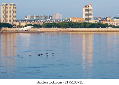 View of China from Russia across the Amur River on a summer morning. A flock of cormorant birds are flying over calm water. City buildings of different styles and purposes. Clear sky. - Shutterstock ID 2175130911