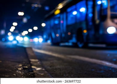 View Of The City At Night, The Bus Driving On The Road. View From The Curb At The Asphalt Level, In Blue Tones