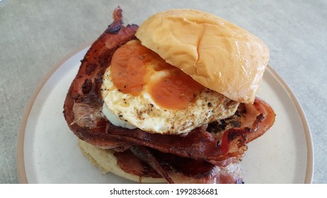 View Of Classic Cafe-style Crispy Bacon And Egg Roll Or Burger With Cheese, Roasted Tomato And Sunny Egg Served On The White Plate With Wooden Table. This Is The Classic And Popular Breakfast Menu. 