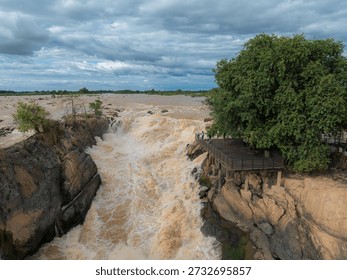 View from cliffside platform overlooking thundering muddy waterfall pouring into a deep gorge surrounded by lush green forest.