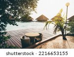 View of a coastal area of a luxury Maldives resort with a selective focus on a wicker cafe table with chairs; a group of typical triangle hotel bungalow-cottages with triangle roofs in the background