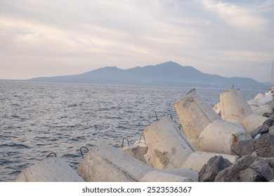 A view of a coastal breakwater with concrete blocks, looking out towards a distant mountain range. The calm blue water reflects the light of the sky. - Powered by Shutterstock