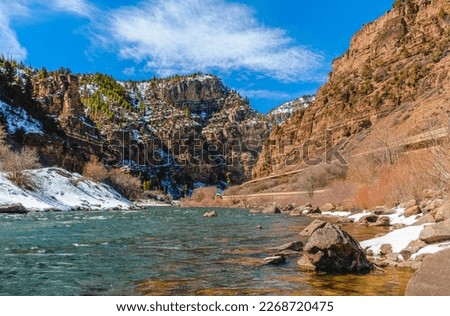 View of Colorado river floating through Glenwood canyon in winter; Rocky Mountains and blue sky in background; distant view of highway 