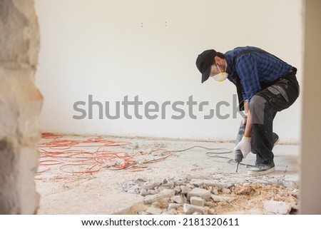 View of a construction worker using a hand-held demolition hammer and breaker to demolish walls to remove old floor tiles during renovation work.