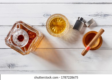 View of a crystal glass and decanter full of golden whisky, and cigar and petrol lighter, shot from above on a distressed white wooden background