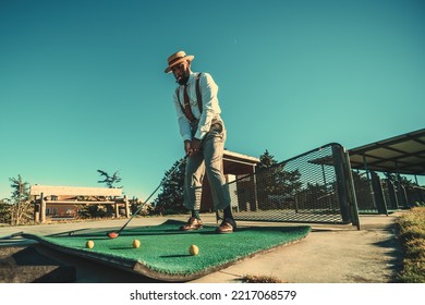 View Of A Dapper African Guy With A Black Beard And A Cigar In His Mouth, In A Straw Hat, He Is Ready To Hit The Ball Using A Club During A Golf Training Warming-up Session On A Training Ground