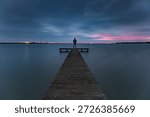 A view of dark clouds during the blue hour above the pier of the Zuiderhaven in Den Oever..

An unrecognizable person stands at the end of a wooden pier in the Zuiderhaven of Den Oever, quietly enjoying the sunrise and the sounds of nature. The rising sun does its best to color the dark clouds, creating a sky rich in contrast.
A moment to pause and be fully present in the here and now. An escape from the hustle and bustle of Western life.