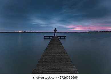 A view of dark clouds during the blue hour above the pier of the Zuiderhaven in Den Oever..

An unrecognizable person stands at the end of a wooden pier in the Zuiderhaven of Den Oever, quietly enjoying the sunrise and the sounds of nature. The rising sun does its best to color the dark clouds, creating a sky rich in contrast.
A moment to pause and be fully present in the here and now. An escape from the hustle and bustle of Western life.