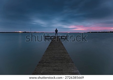 A view of dark clouds during the blue hour above the pier of the Zuiderhaven in Den Oever..

An unrecognizable person stands at the end of a wooden pier in the Zuiderhaven of Den Oever, quietly enjoying the sunrise and the sounds of nature. The rising sun does its best to color the dark clouds, creating a sky rich in contrast.
A moment to pause and be fully present in the here and now. An escape from the hustle and bustle of Western life.