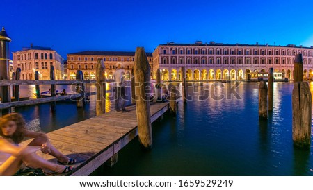 View of the deserted Rialto Market day to night transition timelapse after sunset, San Polo, Venice, Italy viewed from pier with boats and people sitting across the Grand Canal