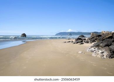 View down a beach flanked by boulders with a massive rock formation deep in the water in the background on a sunny day. 
