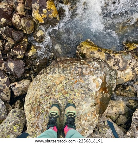 View down on the legs and trekking shoes of a traveler girl standing on alpine stones against the background of a river in the Altai mountains.