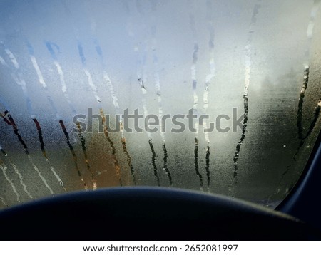A view from the driver's seat looking through a car windshield covered in condensation, fogging and rain streaks. The moisture blurs the outside scenery, creating a soft, abstract effect. 