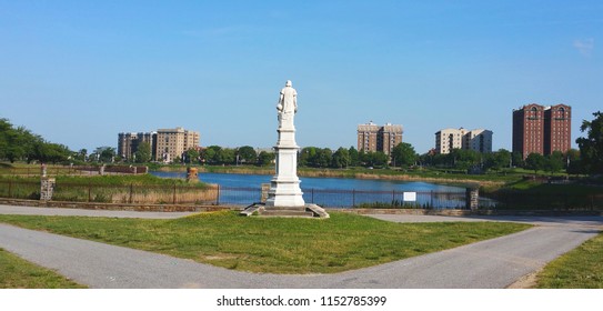 View Of Druid Hill Park  (Baltimore, MD).  Statue Of Christopher Columbus.  View Of Reservoir.  Tall, Residential Buildings Across The Street From The Park.  Clear, Blue Sky.