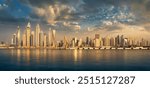 View of the Dubai skyline at sunset from the sea, with the buildings in a row on the horizon reflected in the water. Dramatic clouds and warm sunlit earth colors.