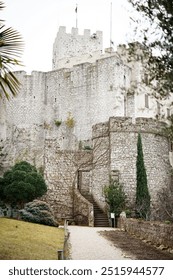 View of Duino Castle, a medieval fortress in Friuli, Italy. With its stone walls, vine-covered stairs, and a tower with a flag, it evokes history and architectural charm in a picturesque setting.