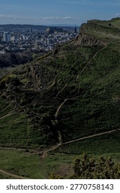 View from Edinburgh looking towards dramatic tree-covered slopes and rocky escarpment with the city visible beyond, Scotland.