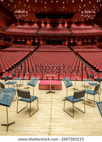 A view of an empty concert hall with rows of red chairs stretching into the distance. In the foreground is a stage with chairs and music stands arranged